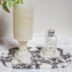 Puede incluir: Una bandeja ovalada decorativa de m&aacute;rmol con un patr&oacute;n blanco y burdeos sostiene un frasco de perfume de vidrio transparente y un jarr&oacute;n de &oacute;nix blanco. El jarr&oacute;n contiene flores artificiales blancas y naranjas. Los art&iacute;culos est&aacute;n sobre un mantel de encaje blanco.