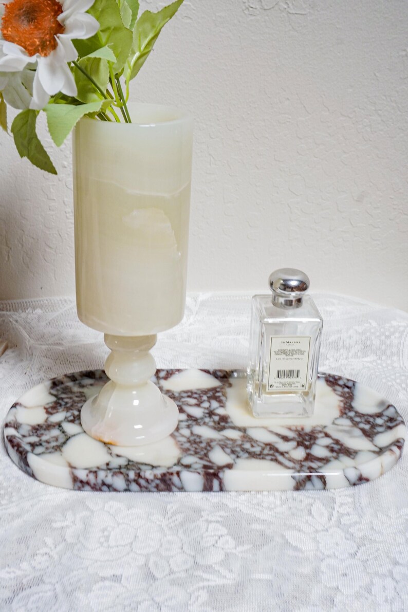 Puede incluir: Una bandeja ovalada decorativa de m&aacute;rmol con un patr&oacute;n blanco y burdeos sostiene un frasco de perfume de vidrio transparente y un jarr&oacute;n de &oacute;nix blanco. El jarr&oacute;n contiene flores artificiales blancas y naranjas. Los art&iacute;culos est&aacute;n sobre un mantel de encaje blanco.