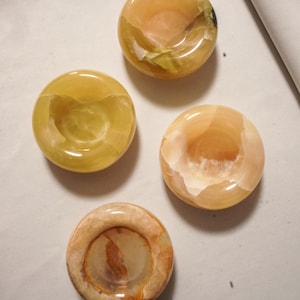 May include: Four circular stone bowls with polished surfaces, each featuring unique natural banding and color variations in shades of amber, honey, yellow, and cream. These small, smooth, concave vessels are arranged in a cluster, showcasing translucent mineral patterns and organic textures throughout the stone material.