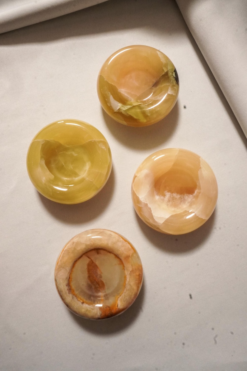 May include: Four circular stone bowls with polished surfaces, each featuring unique natural banding and color variations in shades of amber, honey, yellow, and cream. These small, smooth, concave vessels are arranged in a cluster, showcasing translucent mineral patterns and organic textures throughout the stone material.