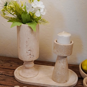 Puede incluir: Un jarr&oacute;n de piedra beige con flores blancas y vegetaci&oacute;n, un candelabro con una vela blanca y una bandeja decorativa. Los objetos est&aacute;n sobre una superficie de madera, creando una est&eacute;tica natural y neutra.