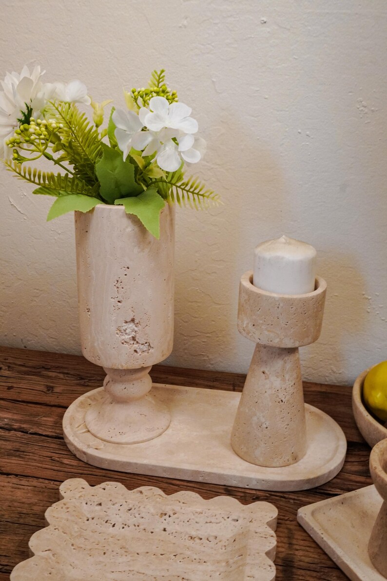 Puede incluir: Un jarr&oacute;n de piedra beige con flores blancas y vegetaci&oacute;n, un candelabro con una vela blanca y una bandeja decorativa. Los objetos est&aacute;n sobre una superficie de madera, creando una est&eacute;tica natural y neutra.