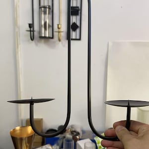 May include: Black metal candle holder with two circular platforms for candles. The holder has a curved, minimalist design. Other decorative items are visible in the background, including a gold-colored vase and glass candle holders.