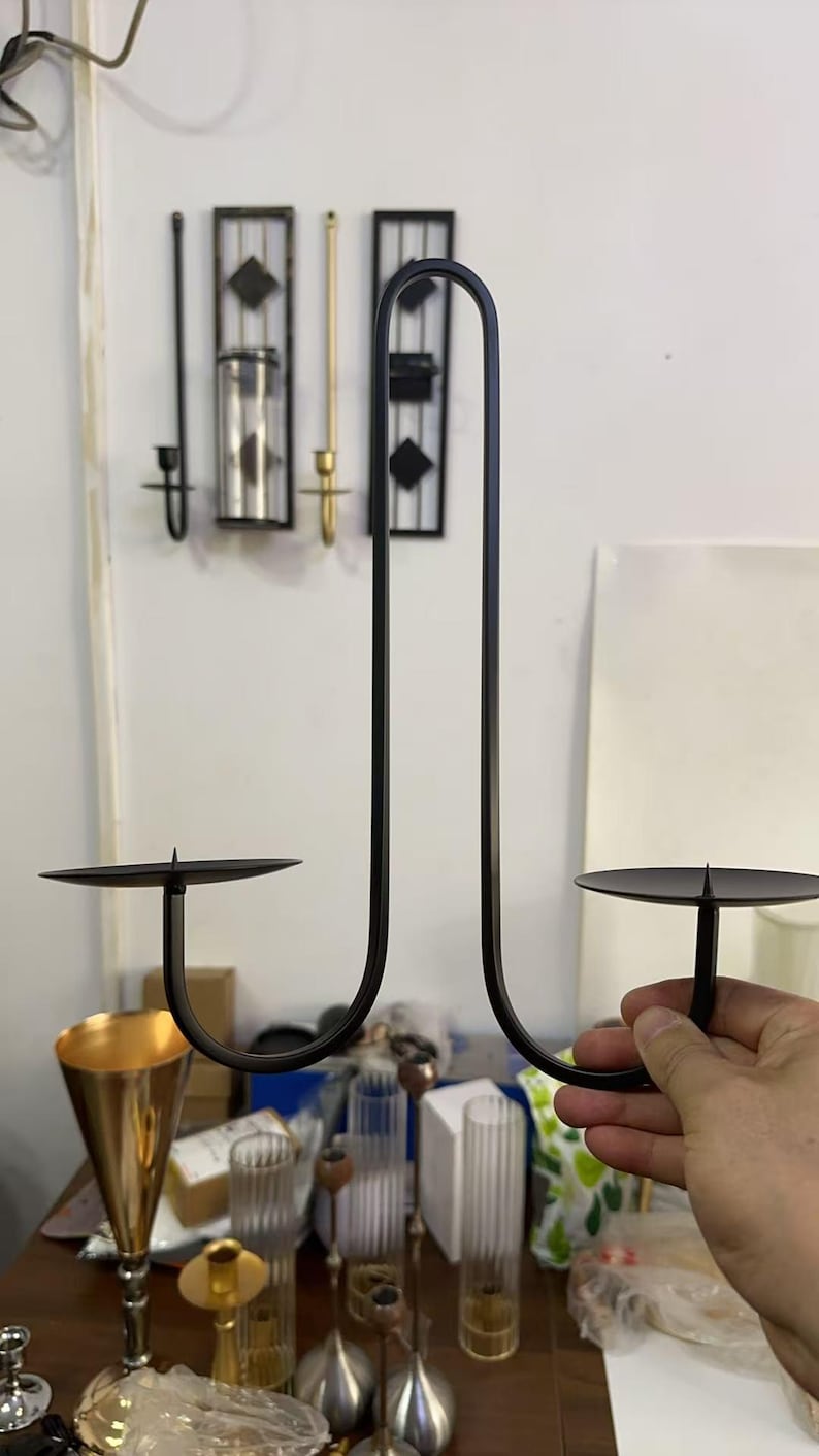 May include: Black metal candle holder with two circular platforms for candles. The holder has a curved, minimalist design. Other decorative items are visible in the background, including a gold-colored vase and glass candle holders.