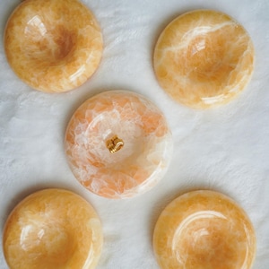 May include: Five circular, polished orange calcite stone bowls with a smooth, glossy finish. Each piece features natural crystalline patterns in varying shades of amber and cream, with a concave center and a small gold-toned metal bail attached.