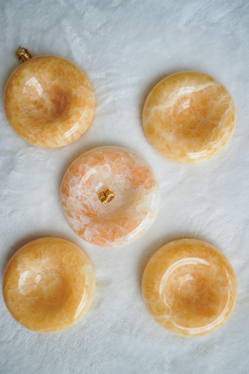 May include: Five circular, polished orange calcite stone bowls with a smooth, glossy finish. Each piece features natural crystalline patterns in varying shades of amber and cream, with a concave center and a small gold-toned metal bail attached.