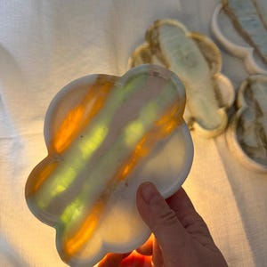 May include: A cloud-shaped tray made of translucent stone featuring horizontal bands of bright yellow, orange, and white. The material allows light to pass through, highlighting the natural veining and color variations within the polished surface.