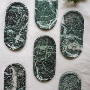 May include: A collection of six oval-shaped marble trays in varying sizes. The trays are a deep green with white veining, creating a natural, patterned appearance. The trays are arranged on a white surface.