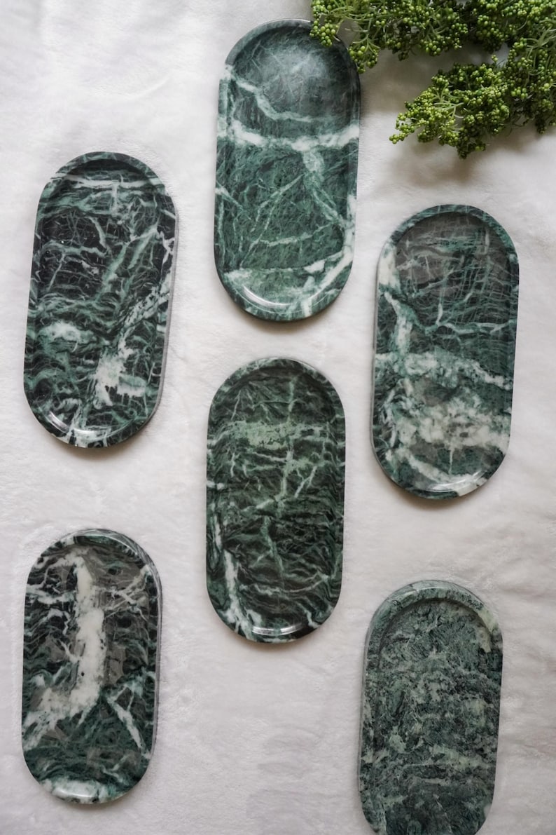 May include: A collection of six oval-shaped marble trays in varying sizes. The trays are a deep green with white veining, creating a natural, patterned appearance. The trays are arranged on a white surface.