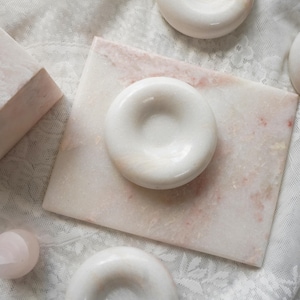 May include: A collection of smooth, polished white marble objects including three circular, donut-shaped pieces with central indentations and one rectangular marble slab featuring subtle pink and white veining throughout the stone surfaces.