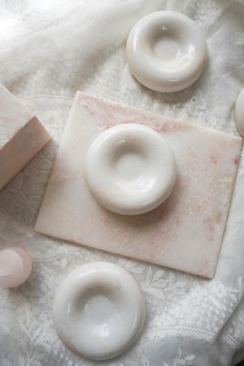 May include: A collection of smooth, polished white marble objects including three circular, donut-shaped pieces with central indentations and one rectangular marble slab featuring subtle pink and white veining throughout the stone surfaces.