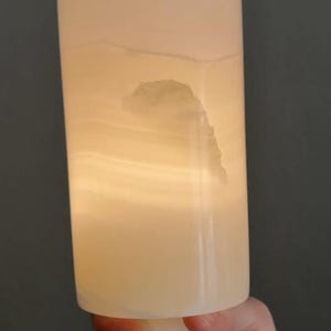 Puede incluir: Una l&aacute;mpara cil&iacute;ndrica de color blanquecino con un interior c&aacute;lido y brillante. La l&aacute;mpara tiene una superficie texturizada y una forma org&aacute;nica &uacute;nica. La base es una forma s&oacute;lida y redondeada. La l&aacute;mpara es sostenida por una persona.