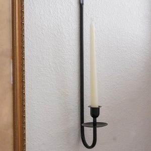 May include: Black metal wall-mounted candle holder with a single ivory-colored candle. The holder has a long, straight stem and a curved base. The candle is inserted into a small, round cup. The holder is mounted on a white wall.
