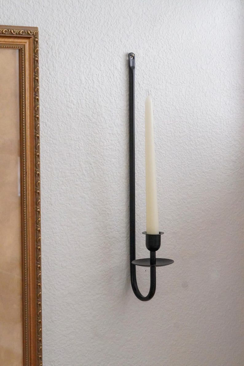 May include: Black metal wall-mounted candle holder with a single ivory-colored candle. The holder has a long, straight stem and a curved base. The candle is inserted into a small, round cup. The holder is mounted on a white wall.