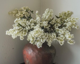 Puede incluir: Un jarrón de cerámica grande, redondeado y de color marrón rojizo oscuro con una superficie texturizada y moteada contiene un arreglo denso y desbordante de flores de lila blancas con pequeñas hojas verdes, creando un despliegue voluminoso.