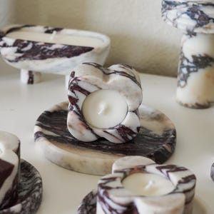 Puede incluir: Una colecci&oacute;n de portavelas con forma de flor hechos de piedra veteada en blanco y morado oscuro. Cada portavelas tiene una abertura circular central con una vela de cera blanca y descansa sobre una bandeja de m&aacute;rmol circular a juego.