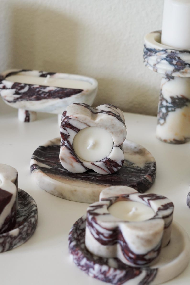 Puede incluir: Una colecci&oacute;n de portavelas con forma de flor hechos de piedra veteada en blanco y morado oscuro. Cada portavelas tiene una abertura circular central con una vela de cera blanca y descansa sobre una bandeja de m&aacute;rmol circular a juego.