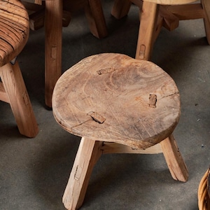 May include: A small, three-legged wooden stool with a round, natural-edge seat. The light brown wood features visible grain, knots, and joinery details where the legs connect to the top. The stool has a weathered, textured surface finish.