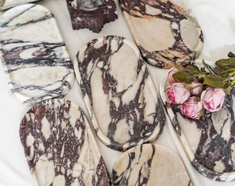 May include: A collection of six oval marble trays featuring cream, white, and deep purple veining. Each tray has a smooth, polished surface and raised edges. A small dark purple stone candle holder and a cluster of pink artificial roses are included.