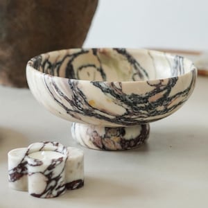 May include: A footed bowl crafted from cream-colored stone with prominent dark brown and black veining throughout. Beside it sits a small, flower-shaped candle holder made from the same marbled stone, featuring a central circular opening for a tea light.