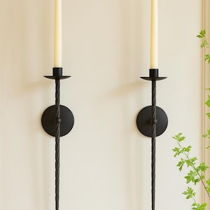 May include: Two black metal wall sconces with a twisted design. Each sconce holds a cream-colored taper candle. The sconces have a circular base and a decorative, elongated stem. The background is a neutral color.