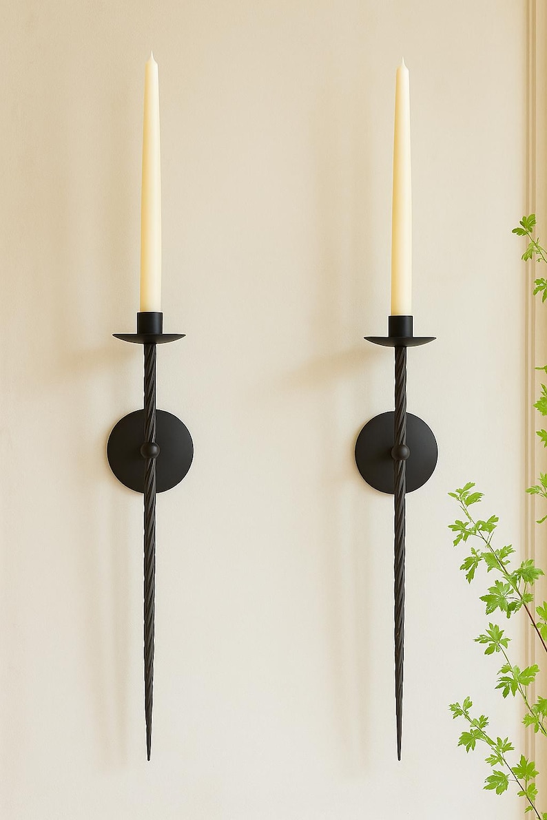 May include: Two black metal wall sconces with a twisted design. Each sconce holds a cream-colored taper candle. The sconces have a circular base and a decorative, elongated stem. The background is a neutral color.