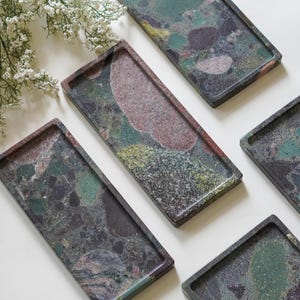 May include: A collection of rectangular trays featuring a multi-colored terrazzo pattern with shades of deep green, dark red, black, and yellow. Each tray has a smooth, flat surface with raised edges, showcasing a unique stone-like composite texture.
