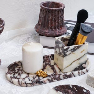 May include: A collection of marble home decor items. Includes a candle, a tray, a pen holder with makeup brushes, and a decorative vase. The marble has a white and burgundy pattern.