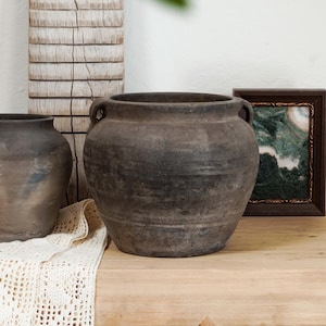 May include: A rustic, dark brown ceramic pot with two handles, displayed on a light wood surface. A smaller pot and a framed picture are also visible. The pot has a wide, rounded shape, suggesting a vintage or antique style.