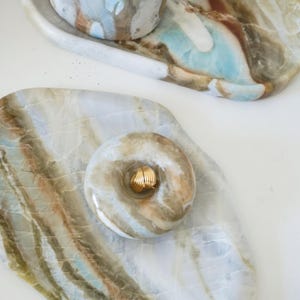 May include: A set of decorative stone trays and a small cup. The trays and cup are made of a stone with a mix of colors including white, brown, and blue. A gold ring sits on top of one of the trays.
