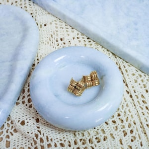 May include: A pair of gold-toned metal stud earrings featuring a woven, textured knot design. The earrings are resting in the center of a small, round, light blue marble dish with a smooth, polished surface and a slightly recessed basin.