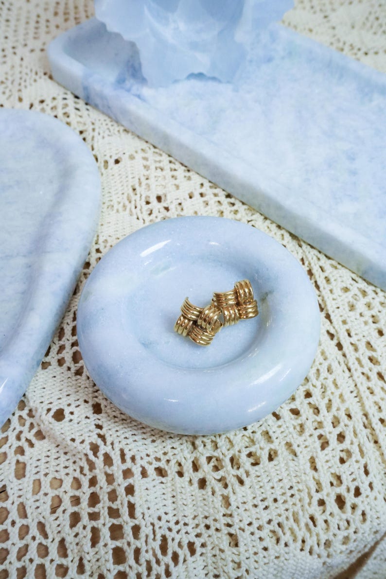 May include: A pair of gold-toned metal stud earrings featuring a woven, textured knot design. The earrings are resting in the center of a small, round, light blue marble dish with a smooth, polished surface and a slightly recessed basin.