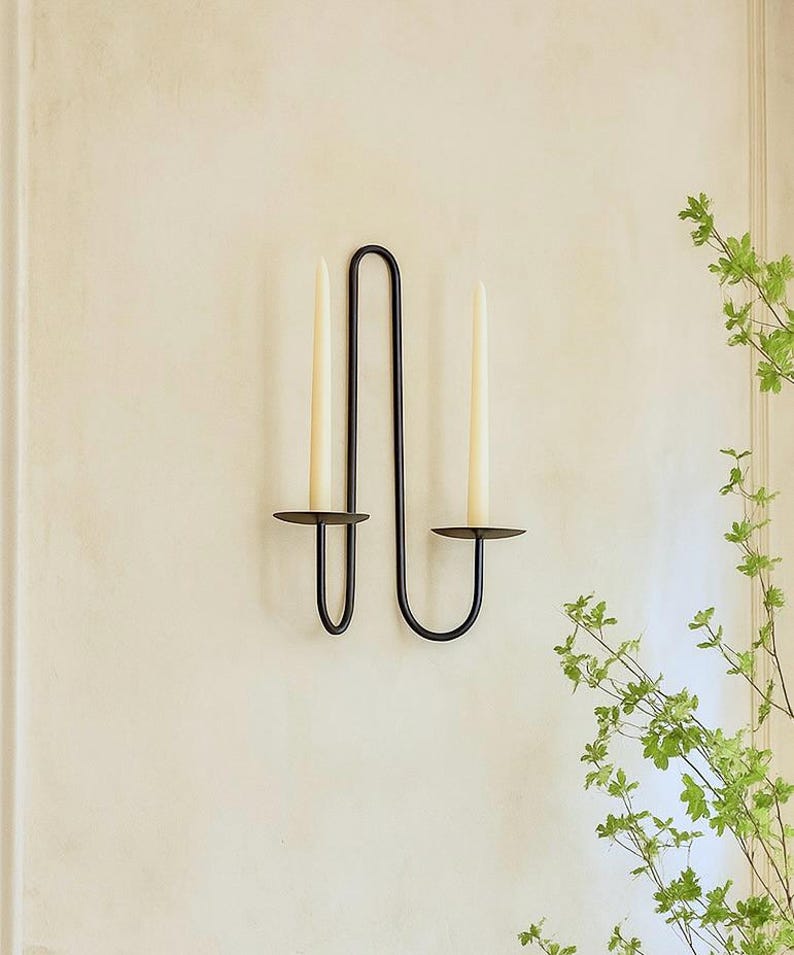 May include: A black metal wall sconce with two candle holders, each holding a tall, cream-colored candle. The sconce has a minimalist design with a curved, vertical structure. Green foliage is visible in the background.