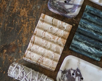 May include: A collection of rectangular soap dishes made from various types of stone, including white and purple marble, green veined stone, and tan patterned stone, alongside a cylindrical bundle of dried sage tied with white string.
