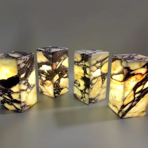 May include: Four rectangular lamps made of translucent stone with a marbled pattern of cream, white, and dark brown veins. Each lamp emits a warm, internal glow, highlighting the natural textures and irregular veining of the stone material.