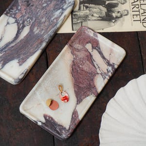 May include: Two rectangular marble trays featuring white and deep burgundy veining. One tray holds a gold-toned oval pendant with a red patterned inlay and a pair of circular earrings in matte gold and terracotta colors. A small gold-toned metal piece sits nearby.