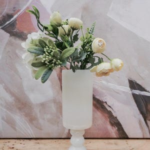 Puede incluir: Un jarr&oacute;n blanco opaco con base de pedestal contiene un ramo de flores artificiales. Las flores incluyen flores blancas y amarillas con follaje verde. El jarr&oacute;n est&aacute; sobre una superficie de madera, con una pintura abstracta borrosa en el fondo.