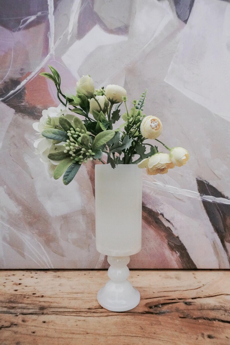 Puede incluir: Un jarr&oacute;n blanco opaco con base de pedestal contiene un ramo de flores artificiales. Las flores incluyen flores blancas y amarillas con follaje verde. El jarr&oacute;n est&aacute; sobre una superficie de madera, con una pintura abstracta borrosa en el fondo.