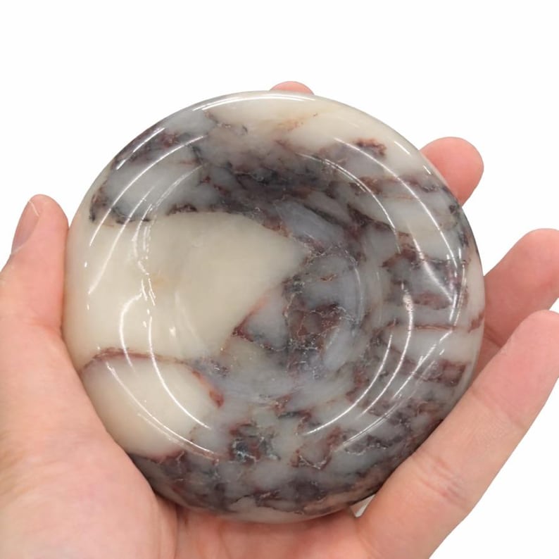 May include: A circular, polished stone dish featuring a smooth, concave surface. The material displays a marbled pattern with creamy white, translucent sections contrasted by deep, dark reddish-brown veins running throughout the mineral structure.