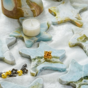 May include: A collection of star-shaped stone coasters and a cylindrical stone holder featuring marbled patterns in shades of light blue, white, and tan. One coaster holds a white tea light candle, another holds a gold knot ring, and a gold bead bracelet.