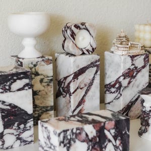 May include: A collection of rectangular marble blocks featuring white, black, and deep purple veining. The blocks serve as pedestals for a white chalice, a flower-shaped candle holder, and a miniature architectural model labeled ROMAS PIETRO.