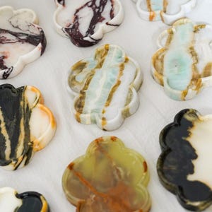 May include: A collection of flower-shaped stone trays featuring various natural patterns and colors, including white with dark veining, translucent green with brown streaks, and black with cream accents, arranged in a group on a white surface.