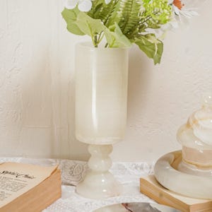 Puede incluir: Un jarr&oacute;n alto de &oacute;nix blanquecino con flores blancas y follaje verde. El jarr&oacute;n tiene un cuerpo cil&iacute;ndrico y una base de pedestal. Tambi&eacute;n se ven otros objetos de &oacute;nix, como un plato y una pieza decorativa.