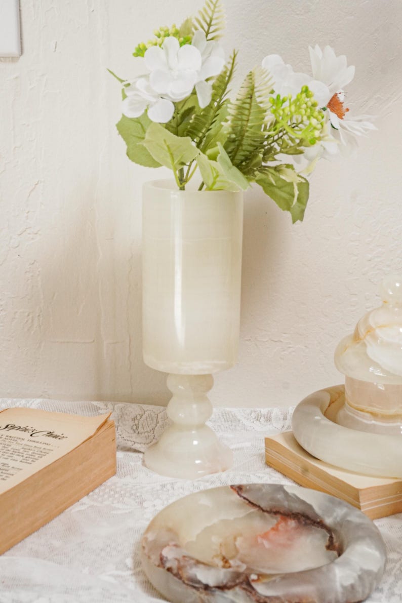 Puede incluir: Un jarr&oacute;n alto de &oacute;nix blanquecino con flores blancas y follaje verde. El jarr&oacute;n tiene un cuerpo cil&iacute;ndrico y una base de pedestal. Tambi&eacute;n se ven otros objetos de &oacute;nix, como un plato y una pieza decorativa.