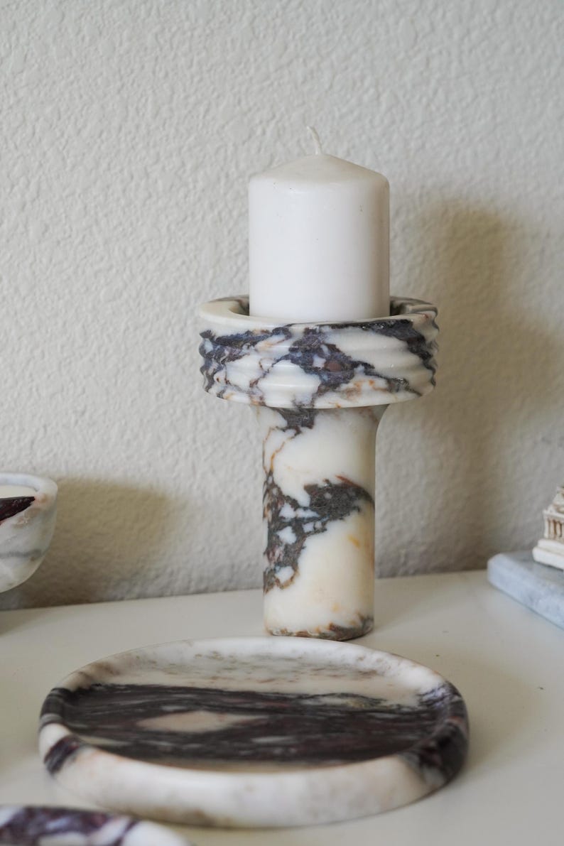 Puede incluir: Un portavelas cil&iacute;ndrico de m&aacute;rmol con un patr&oacute;n veteado en blanco, negro y marr&oacute;n, que presenta un borde superior ancho y acanalado. Una vela de pilar blanca s&oacute;lida est&aacute; insertada en el centro del soporte, situado detr&aacute;s de una bandeja de m&aacute;rmol.