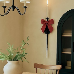 May include: A dining room scene featuring a black chandelier with cream-colored shades, a vase with greenery, and a wooden table. A black wall sconce with a red bow holds a lit candle, adding a warm ambiance.