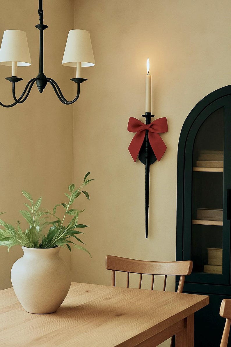 May include: A dining room scene featuring a black chandelier with cream-colored shades, a vase with greenery, and a wooden table. A black wall sconce with a red bow holds a lit candle, adding a warm ambiance.