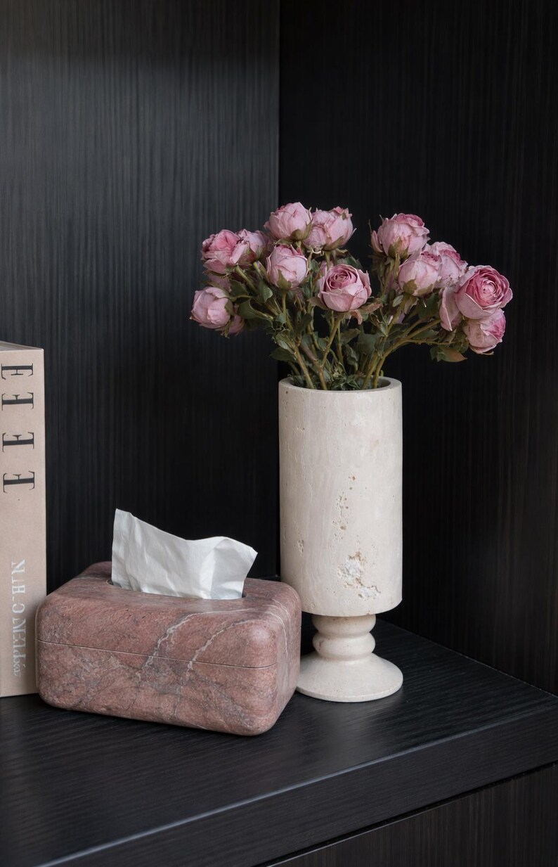 Puede incluir: Una composici&oacute;n decorativa con una caja de pa&ntilde;uelos de m&aacute;rmol rosa, un jarr&oacute;n crema lleno de rosas rosas y un libro. Los objetos se colocan sobre un fondo oscuro y texturizado, creando una escena elegante.