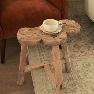 May include: A rustic, three-legged wooden stool with a unique, organic shape. A white teacup and saucer sit atop the stool. The stool is made of light-colored wood and has visible grain and knots. The background includes a rust-colored armchair and a patterned rug.