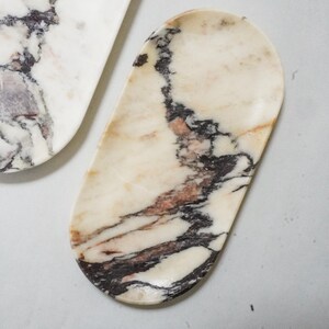 May include: Two marble trays with a natural stone pattern. The trays have a cream-colored base with dark brown and reddish-brown veining. One tray is oval-shaped, and the other is a long, rectangular shape.